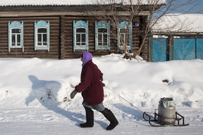 An elderly woman pulling a metal container with water past a snow-covered wooden house with carved window frames in the village of Kondratyevo, Omsk Region, February 17, 2017. Photo courtesy of Dmitry Feoktistov/TASS Россия. Омская область. 19 февраля 2017. Жительница села Кондратьево в Муромцевском районе Омской области. Дмитрий Феоктистов/ТАСС
